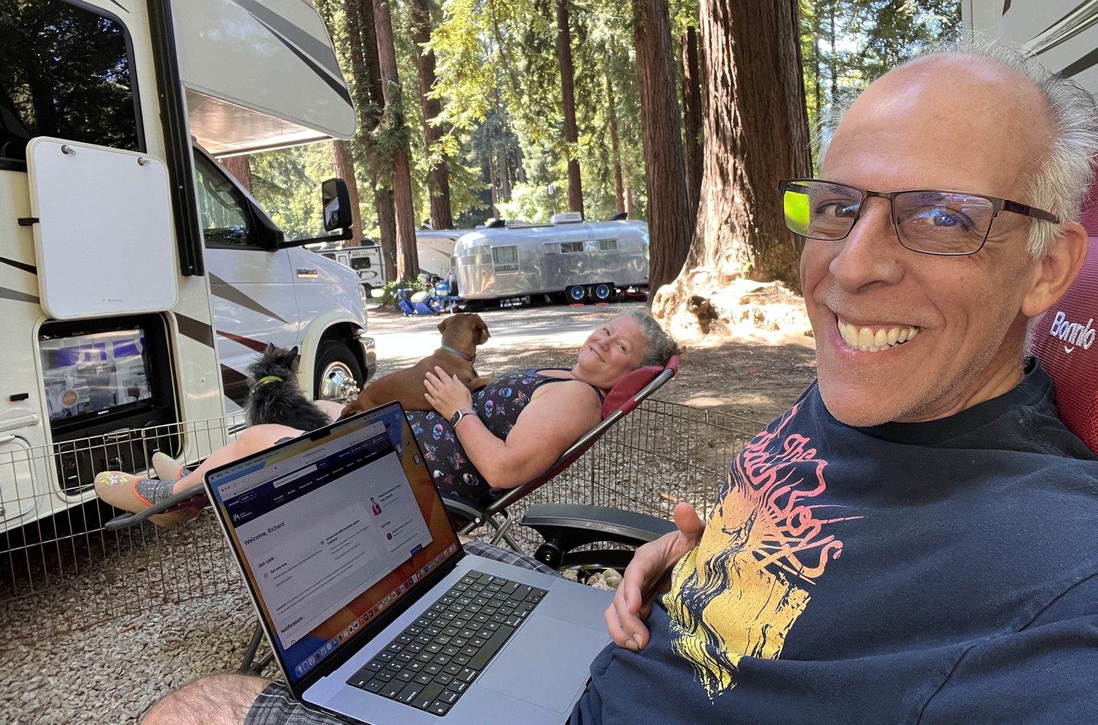 Richard working on his laptop at an RV campsite in the redwoods, with his wife and dogs.
